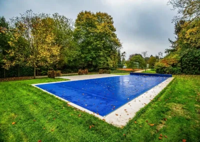 Piscine couverte d'une bâche bleue entourée de pelouse verte et d'arbres aux feuilles jaunissantes.