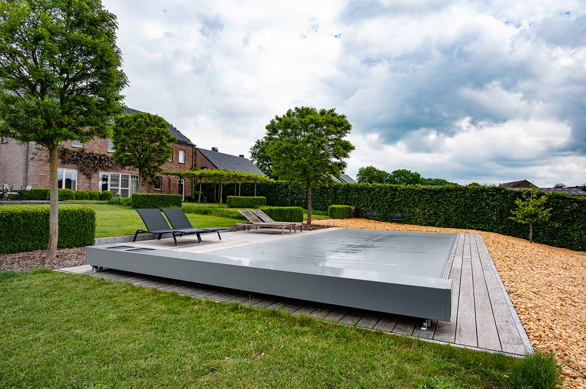 Terrasse avec piscine couverte grise, entourée de transats noirs, pelouse verte, arbres et haies, devant une maison en briques rouges.