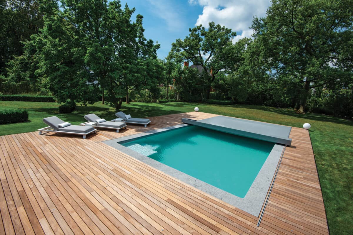 Terrasse en bois avec piscine rectangulaire turquoise, entourée de chaises longues grises et de pelouse verte, sous un ciel bleu partiellement nuageux.