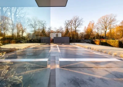 Allée en béton avec des reflets de verre, bordée de haies et d'arbres nus sous un ciel clair et ensoleillé.