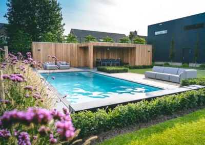 Piscine entourée de haies et de fleurs violettes, avec un salon de jardin en bois et des canapés gris sur la terrasse.