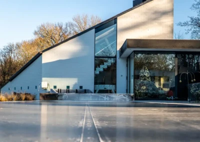 Maison moderne avec façade blanche et grandes fenêtres, entourée d'arbres dénudés, et terrasse en béton au premier plan.