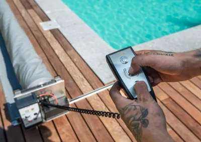 Des mains tiennent une télécommande noire près d'une piscine, avec un enrouleur de couverture gris sur un sol en bois.