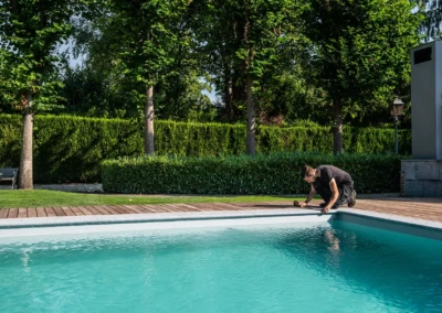 Une personne ajuste le bord d'une piscine entourée d'une terrasse en bois, avec des arbres et des haies en arrière-plan.