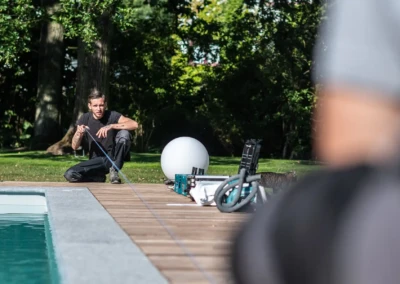 Un homme agenouillé mesure le bord d'une piscine avec un ruban à mesurer, entouré d'arbres et d'équipements divers.