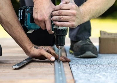 Deux mains installent une vis avec une perceuse sans fil sur un rail métallique posé sur une surface en bois et tapis gris.
