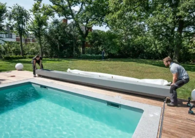 Deux personnes installent une couverture de piscine blanche sur une terrasse en bois, entourée d'un jardin verdoyant.