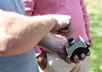 Deux personnes manipulant un boîtier métallique avec une clé, une portant un t-shirt gris et l'autre un polo rose, en extérieur.