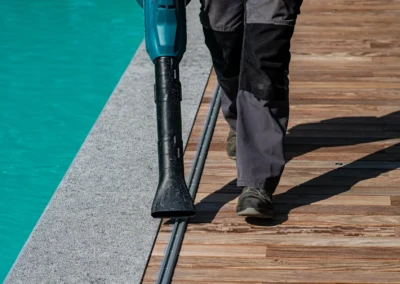 Un homme en pantalon noir utilise un souffleur de feuilles bleu sur une terrasse en bois près d'une piscine turquoise.