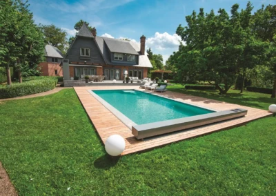 Grande maison en briques avec terrasse en bois et piscine rectangulaire, entourée de pelouse verte et d'arbres sous un ciel bleu.