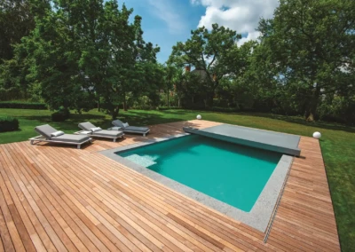 Terrasse en bois avec quatre transats gris autour d'une piscine rectangulaire, entourée de pelouse et d'arbres verts.