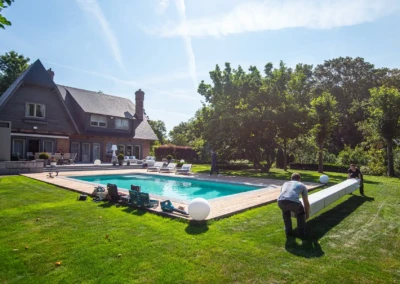 Deux personnes installent une couverture de piscine blanche près d'une maison en briques avec un grand jardin verdoyant.