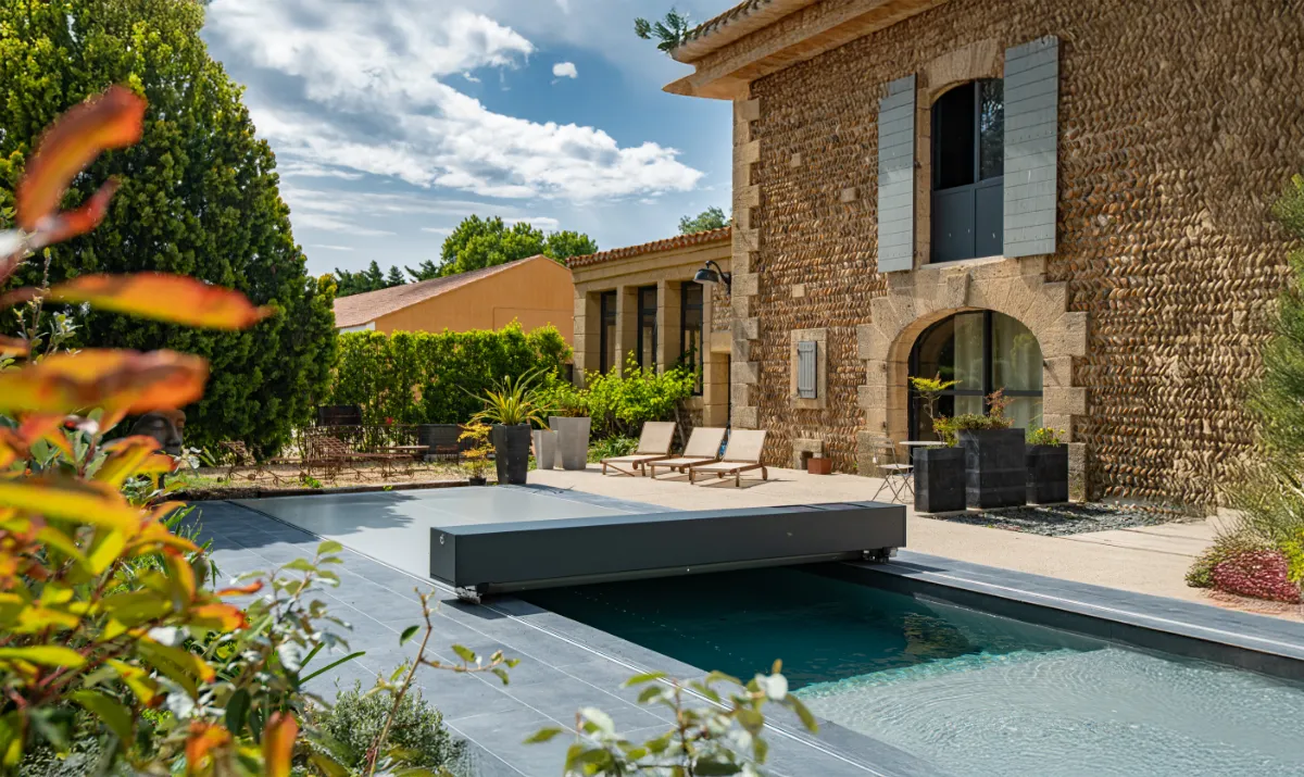 Une piscine avec couverture grise automatique est entourée de plantes et de chaises longues, près d'une maison en pierre.