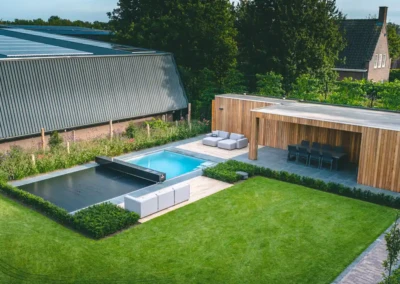 Une piscine avec couverture grise est entourée de mobilier de jardin et d'une terrasse en bois, avec pelouse verte et haies soignées.
