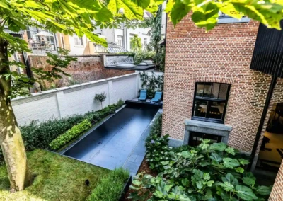 Cour arrière avec piscine couverte noire, entourée de murs en briques rouges et de végétation, deux chaises longues bleues visibles.