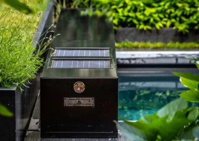 Boîtier noir avec panneaux solaires à côté d'une piscine, entouré de plantes vertes et de fleurs violettes.