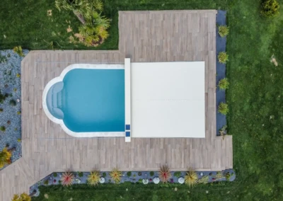 Vue aérienne d'une piscine rectangulaire avec couverture blanche, entourée de terrasse en bois et de pelouse verte.
