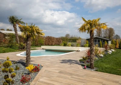 Jardin avec piscine entourée de palmiers, terrasse en bois et cabanon en arrière-plan sous un ciel nuageux.