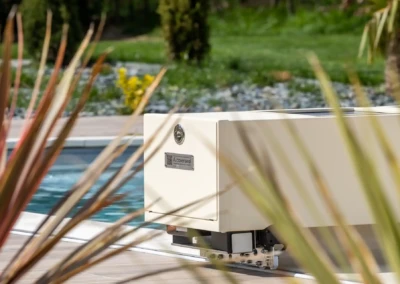 Un boîtier beige de couverture de piscine est placé à côté d'une piscine entourée de verdure et de plantes.