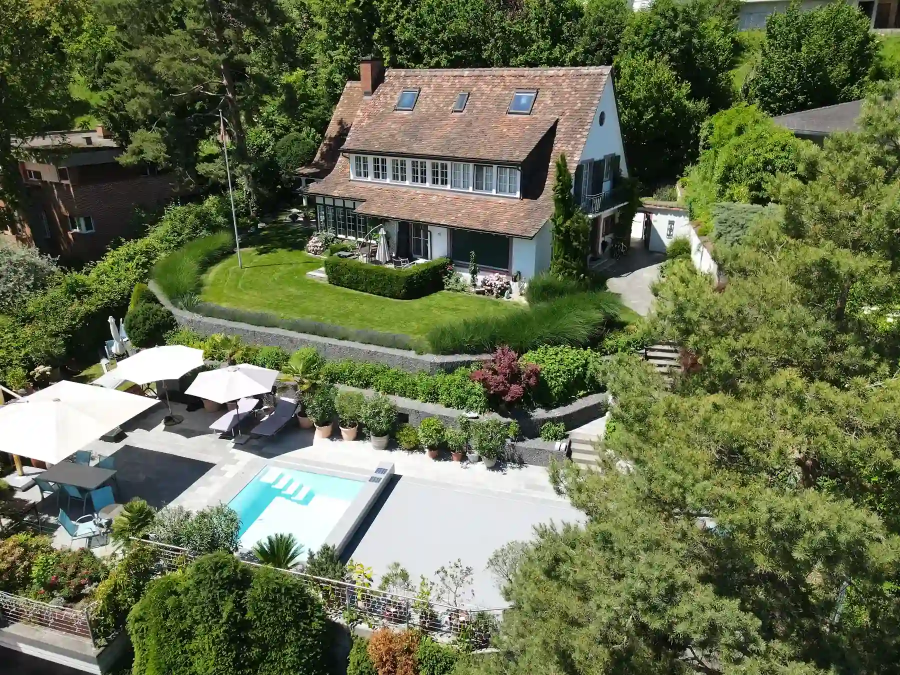 Maison avec toit en tuiles entourée de verdure, piscine et terrasse avec parasols blancs au premier plan.