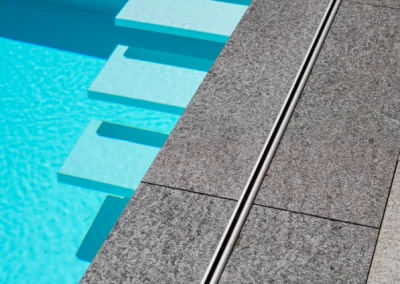 Des dalles grises en pierre entourent une piscine d'eau bleue avec des marches immergées.