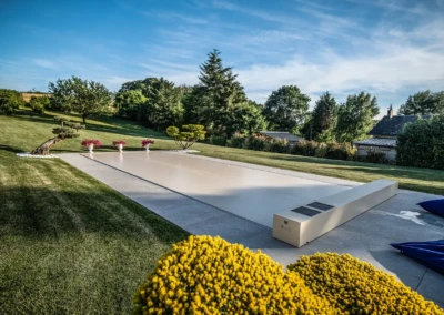 Couverture de piscine beige entourée de pelouse verte, avec des pots de fleurs roses et des arbres en arrière-plan.