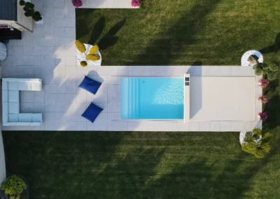 Vue aérienne d'une piscine rectangulaire, entourée de dalles blanches, avec des coussins bleus et des arbustes décoratifs sur pelouse verte.