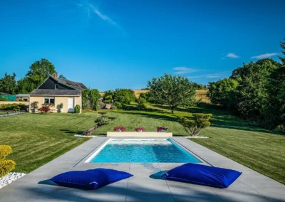 Maison beige avec toit noir, piscine entourée de coussins bleus, pelouse verte et arbres en arrière-plan sous ciel bleu.