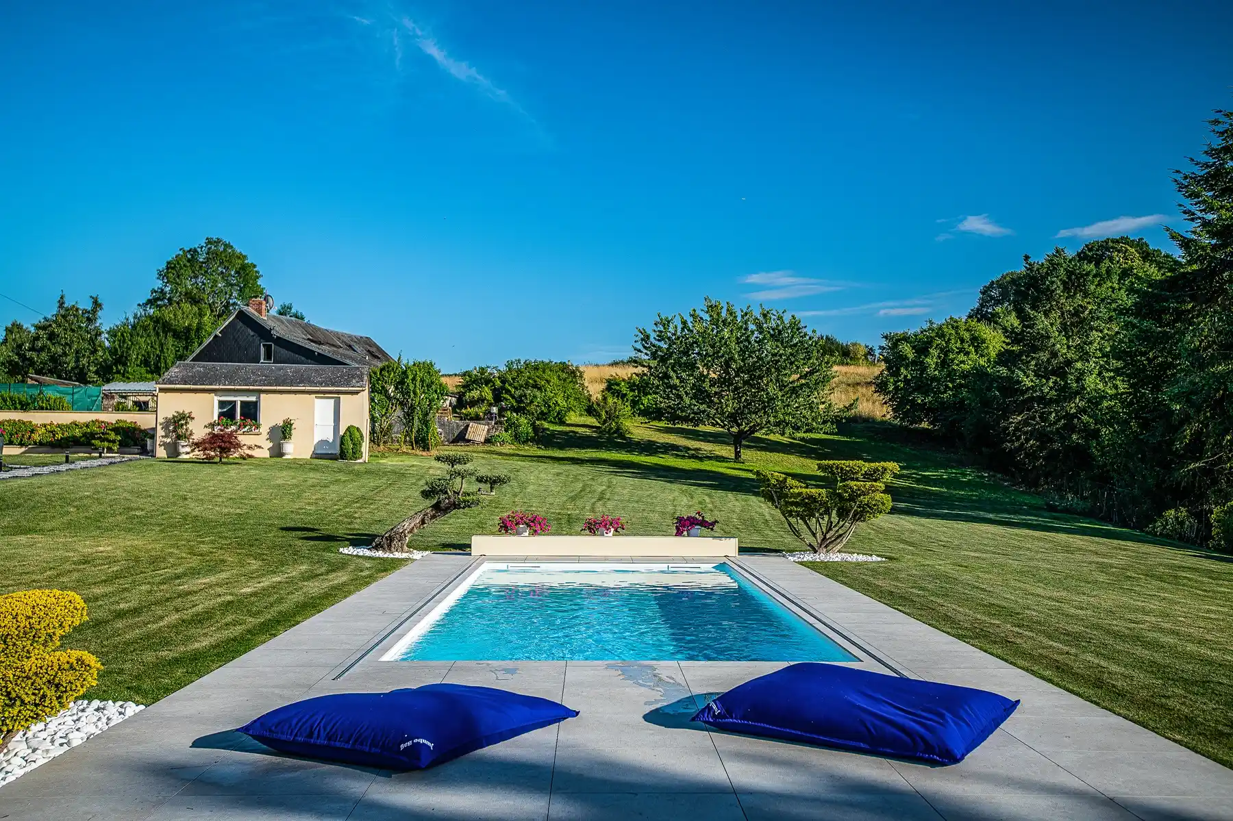 Maison beige avec toit noir, piscine entourée de coussins bleus, pelouse verte et arbres en arrière-plan sous ciel bleu.