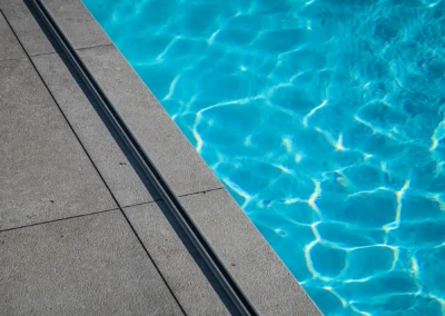 Surface d'une piscine avec eau bleue et reflets lumineux, bordée par un carrelage gris clair.