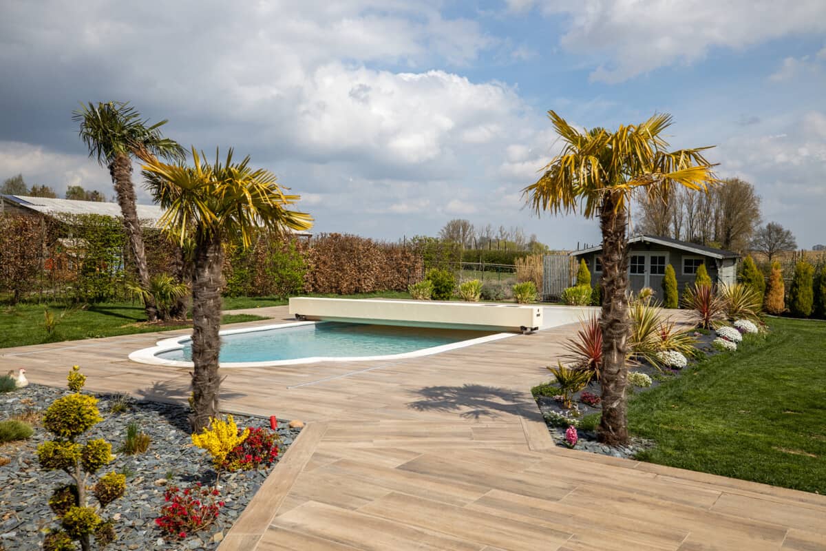 Une piscine entourée de palmiers et de plantes colorées, avec un abri de jardin en arrière-plan sous un ciel partiellement nuageux.