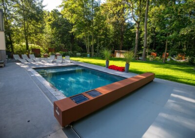 Piscine rectangulaire entourée de chaises longues blanches, bordée par une terrasse en béton et une pelouse verte avec des arbres en arrière-plan.