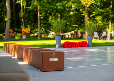 Une longue boîte en métal rouille est posée sur une terrasse en béton devant un jardin verdoyant avec des plantes en pot.