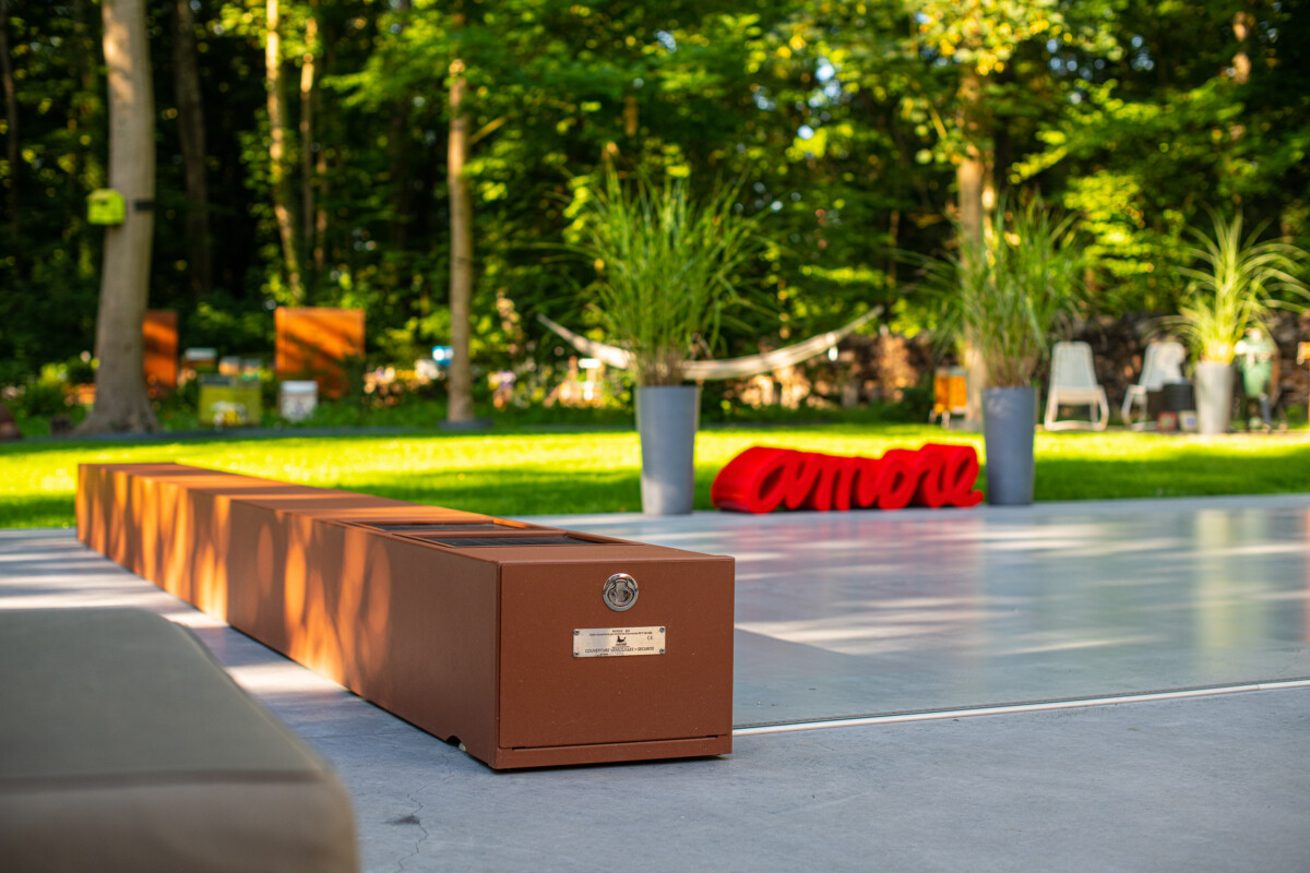 Une longue boîte en métal rouille est posée sur une terrasse en béton devant un jardin verdoyant avec des plantes en pot.