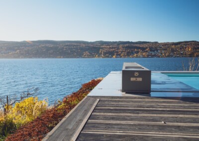 Terrasse en bois avec vue sur un lac bleu, entourée de collines boisées sous un ciel dégagé, avec un objet rectangulaire gris au bord de l'eau.