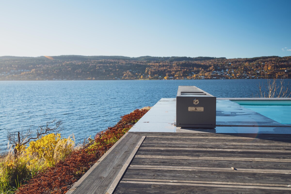 Terrasse en bois avec vue sur un lac bleu, entourée de collines boisées sous un ciel dégagé, avec un objet rectangulaire gris au bord de l'eau.