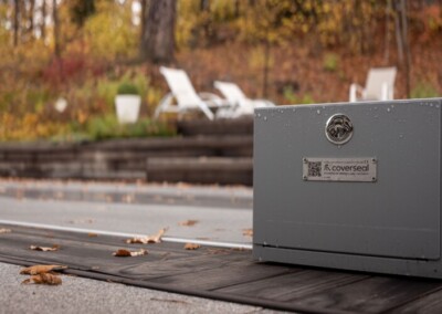Boîtier métallique gris avec logo, entouré de feuilles mortes, devant des chaises longues blanches sur une terrasse en bois.