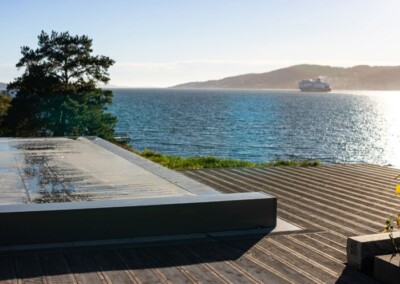 Une couverture de piscine grise sur une terrasse en bois surplombe un paysage de mer avec un bateau à l'horizon.