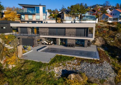 Grande maison moderne à deux étages avec façade en verre et pierre, entourée de végétation automnale et de maisons voisines, sous un ciel bleu clair.
