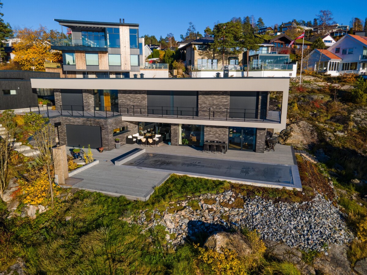 Grande maison moderne à deux étages avec façade en verre et pierre, entourée de végétation automnale et de maisons voisines, sous un ciel bleu clair.