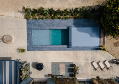 Vue aérienne d'une piscine rectangulaire avec couverture grise, entourée de dalles grises et d'un jardin aménagé avec des chaises longues.