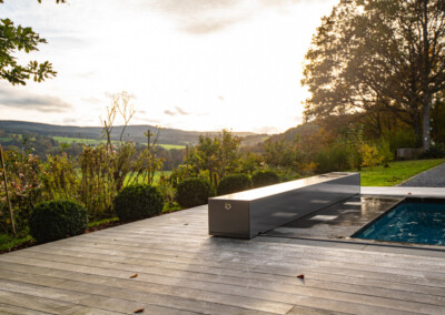 Terrasse en bois avec piscine couverte, entourée de buissons verts, sous un ciel nuageux avec vue sur des collines verdoyantes.