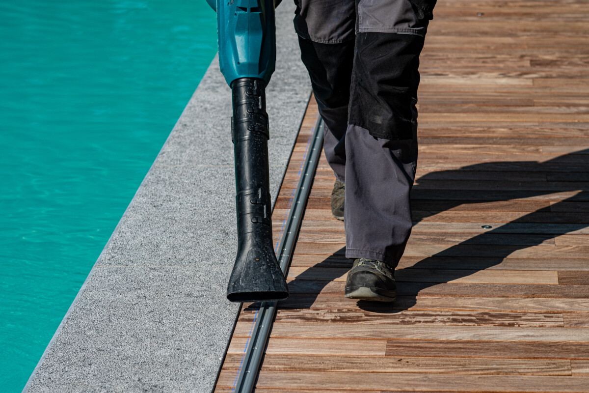 Un homme en pantalon gris utilise un souffleur noir sur une terrasse en bois près d'une piscine turquoise.