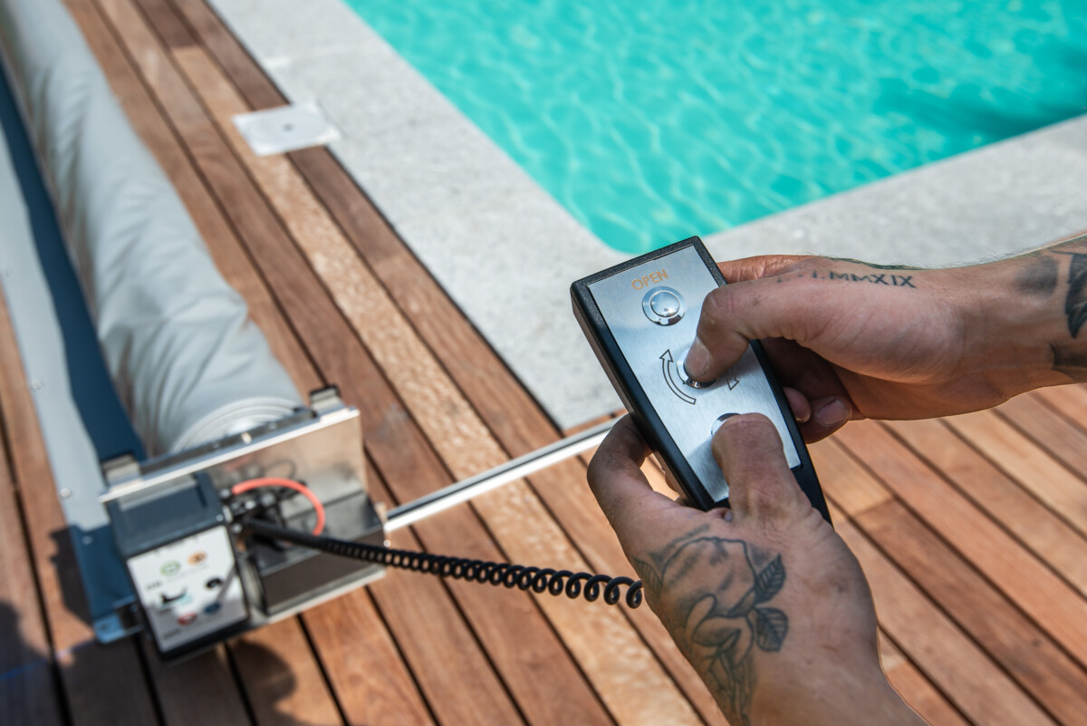 Une télécommande noire est utilisée pour ouvrir une couverture de piscine grise, située sur une terrasse en bois près de l'eau turquoise.