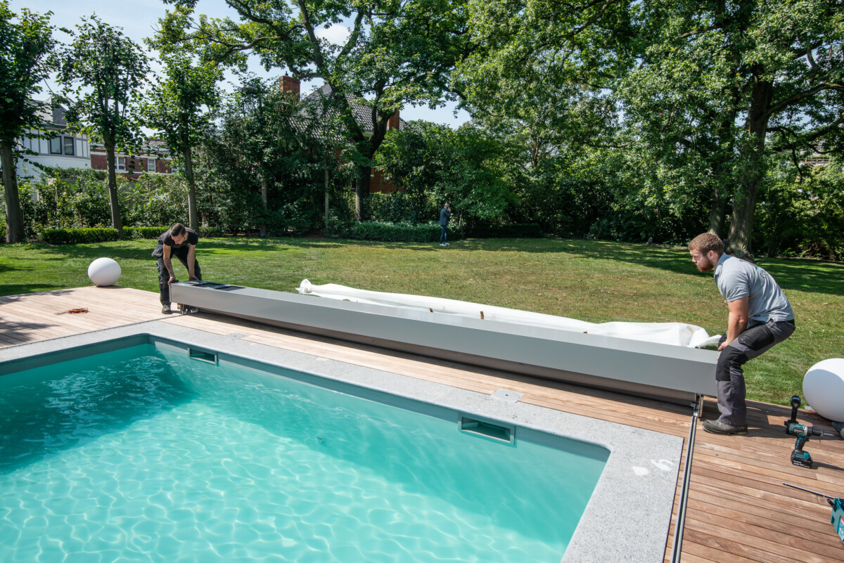 Deux personnes installent un volet roulant blanc sur une piscine entourée de bois, avec un jardin verdoyant en arrière-plan.
