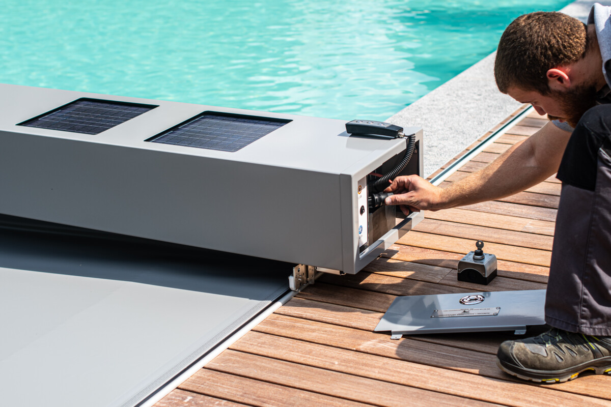 Un homme ajuste un boîtier gris avec des panneaux solaires à côté d'une piscine, sur une terrasse en bois.