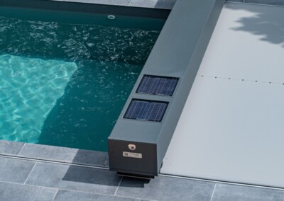 Une piscine avec une couverture grise motorisée et des panneaux solaires, entourée de dalles grises et de végétation verte.