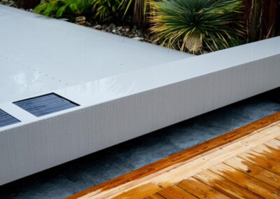 Une couverture de piscine grise avec des panneaux solaires est entourée de plantes vertes et d'une terrasse en bois.