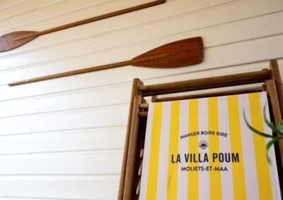Deux pagaies en bois sont accrochées sur un mur blanc, à côté d'un transat jaune et blanc marqué "La Villa Poum".