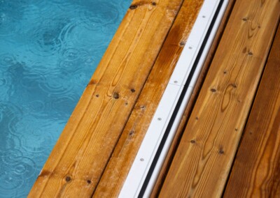 Planches en bois clair bordent une piscine bleue avec des gouttes de pluie formant des cercles à la surface de l'eau.